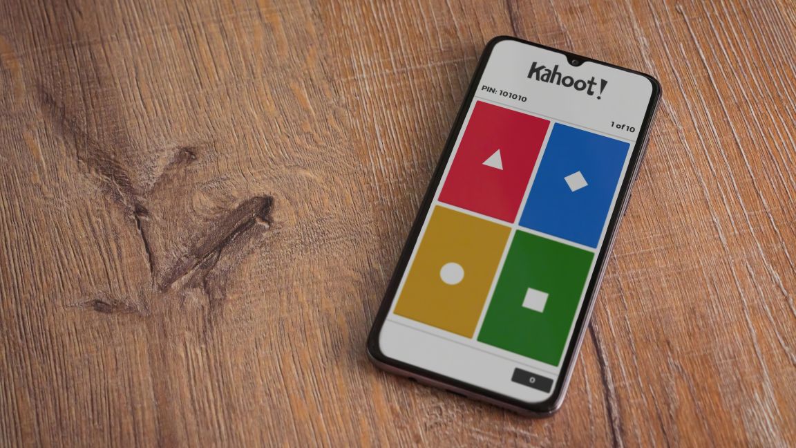 Startbildschirm der Kahoot!‑App mit Logo auf dem Display eines schwarzen Smartphones vor hölzernem Hintergrund.